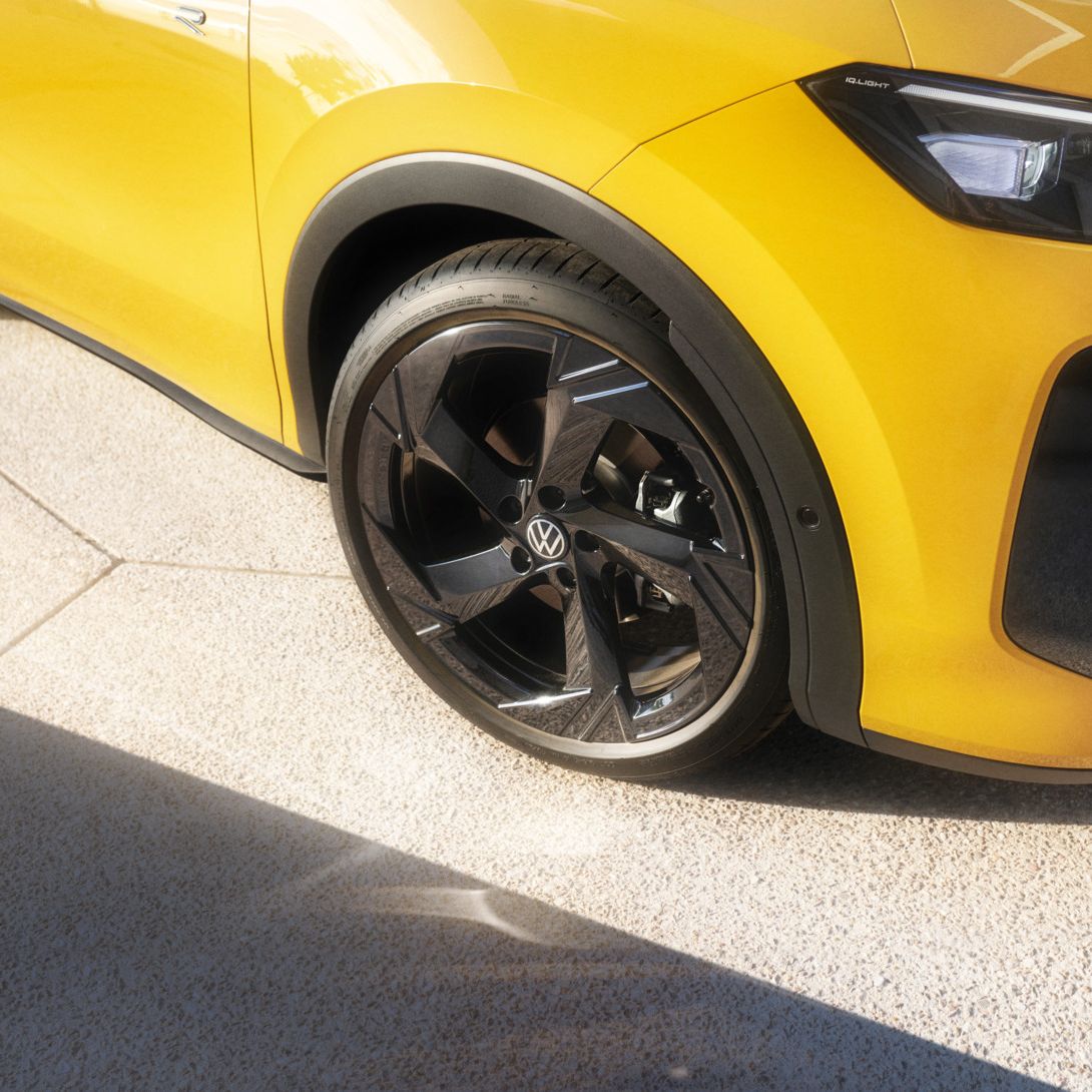 Das Bild zeigt einen Teil eines gelben T-Roc aus der Seitenansicht. Der Fokus liegt auf dem schwarzen Vorderrad und der Kotflügel. Das Auto steht auf hellen, rauen Bodenplatten.