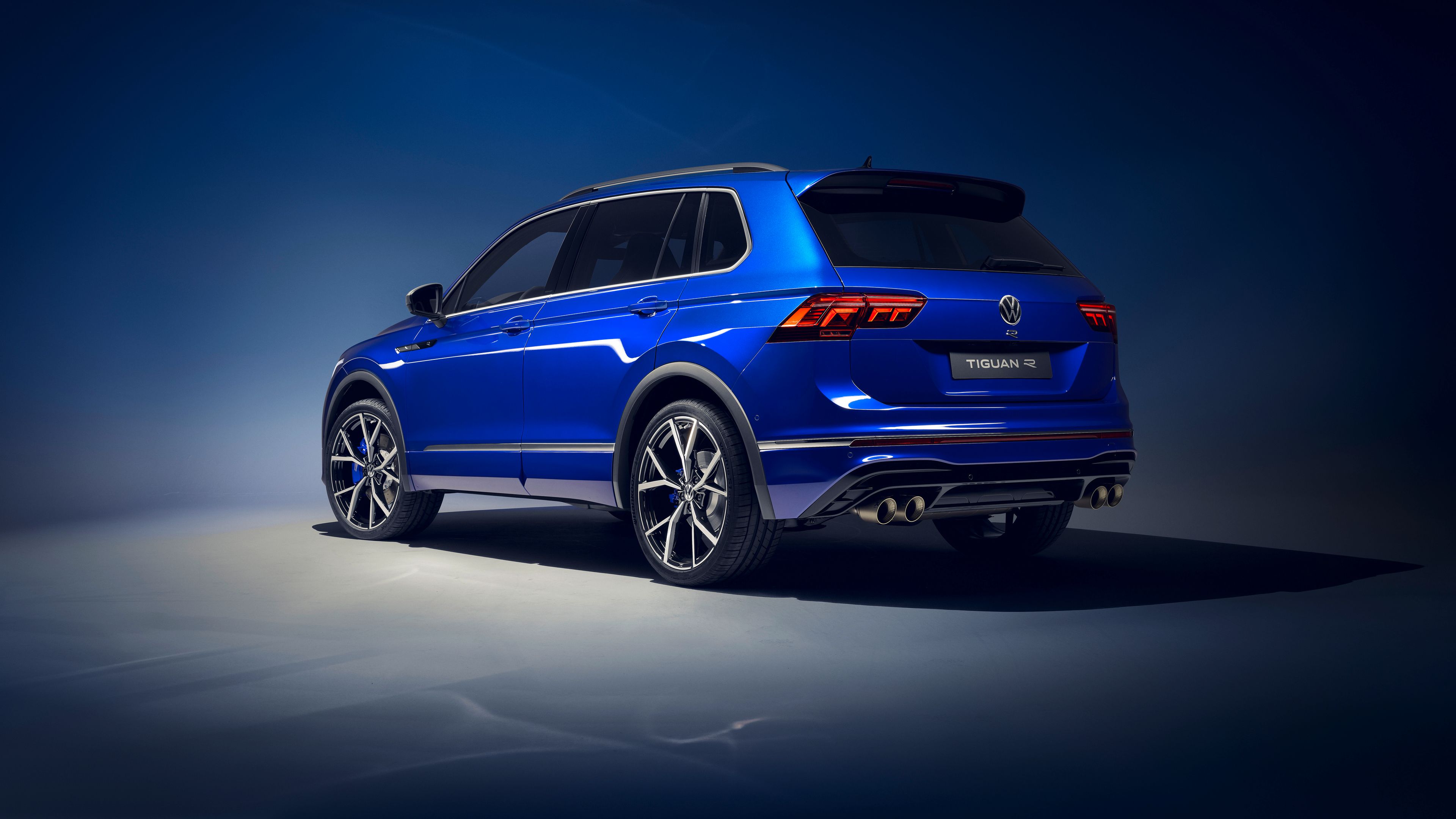 Ein blauer Volkswagen Tiguan R wird von hinten und seitlich gezeigt. Das Auto steht vor einem dunklen Hintergrund und hat moderne LED-Rückleuchten. Die Sportversion hat zwei große Auspuffrohre auf jeder Seite hinten.