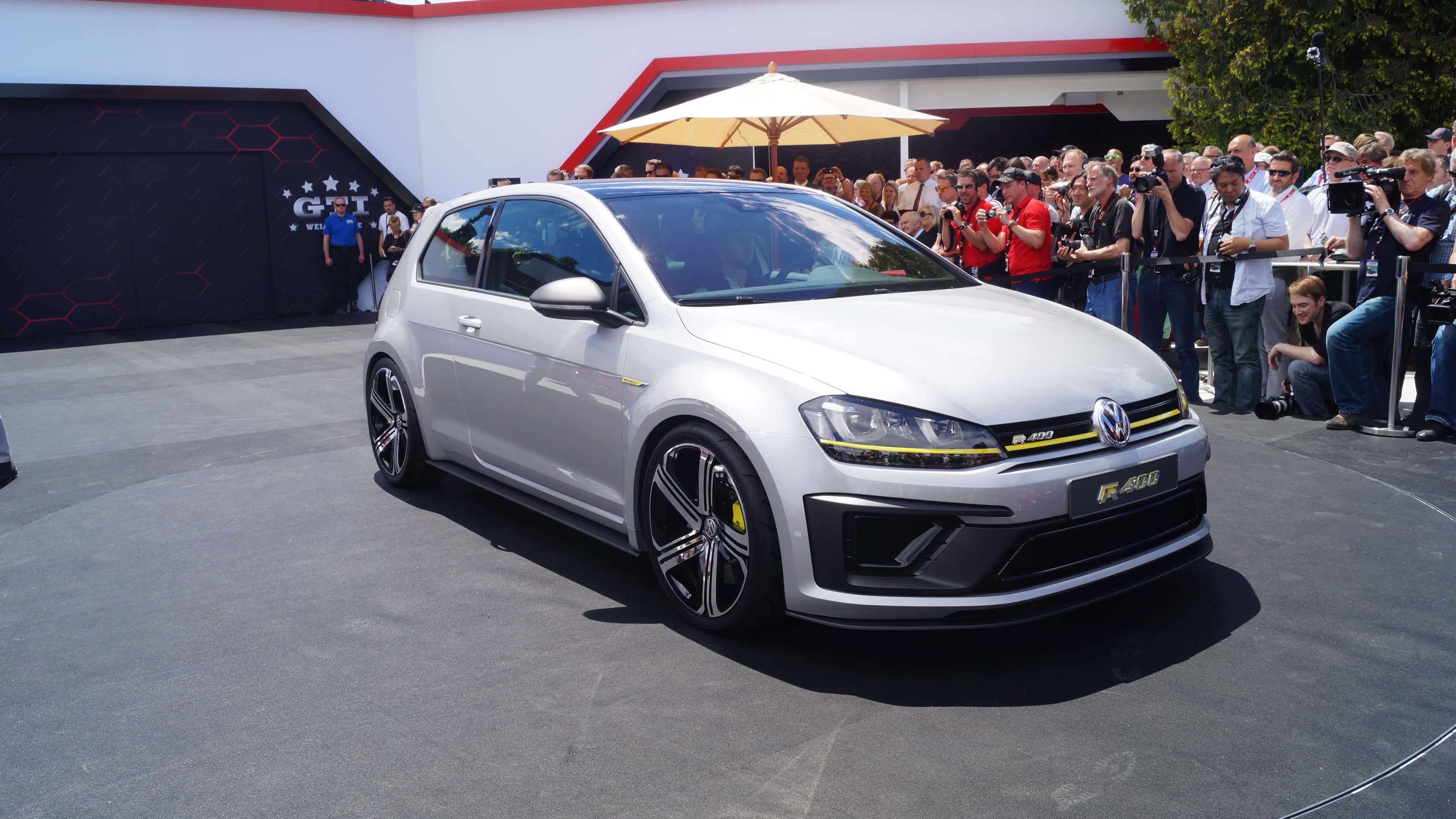 Das Bild zeigt einen silberfarbenen Volkswagen Golf R, der bei einem Event präsentiert wird. Hintergrund gibt es eine Gruppe von Menschen, die das Auto betrachten.