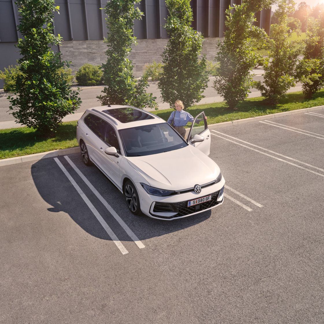 Eine Frau öffnet auf einem begrünten Parkplatz die Fahrertür eines weißen VW Passat R-Line und steigt ein.