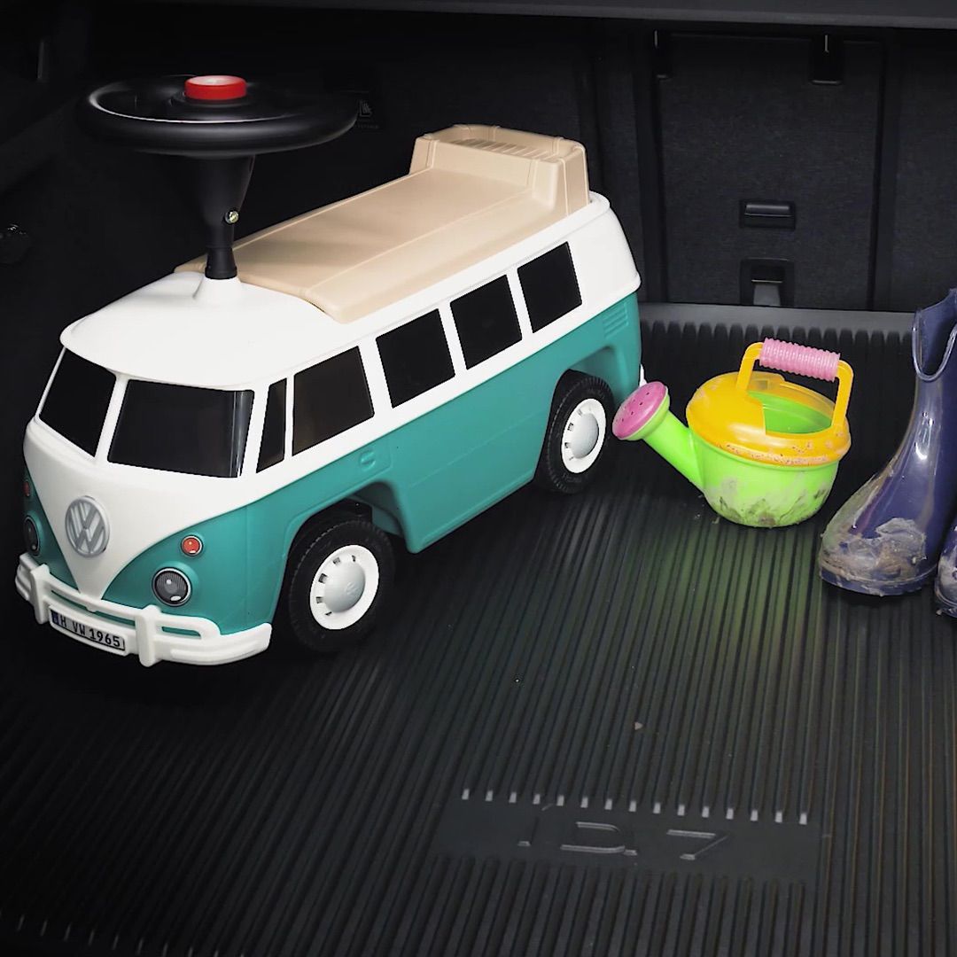 Spielzeug-Lkw in Form eines VW-Busses steht neben einer Gießkanne und einem Paar blauer Gummistiefel. Alle Objekte befinden sich auf einer schwarzen, gerillten Oberfläche.