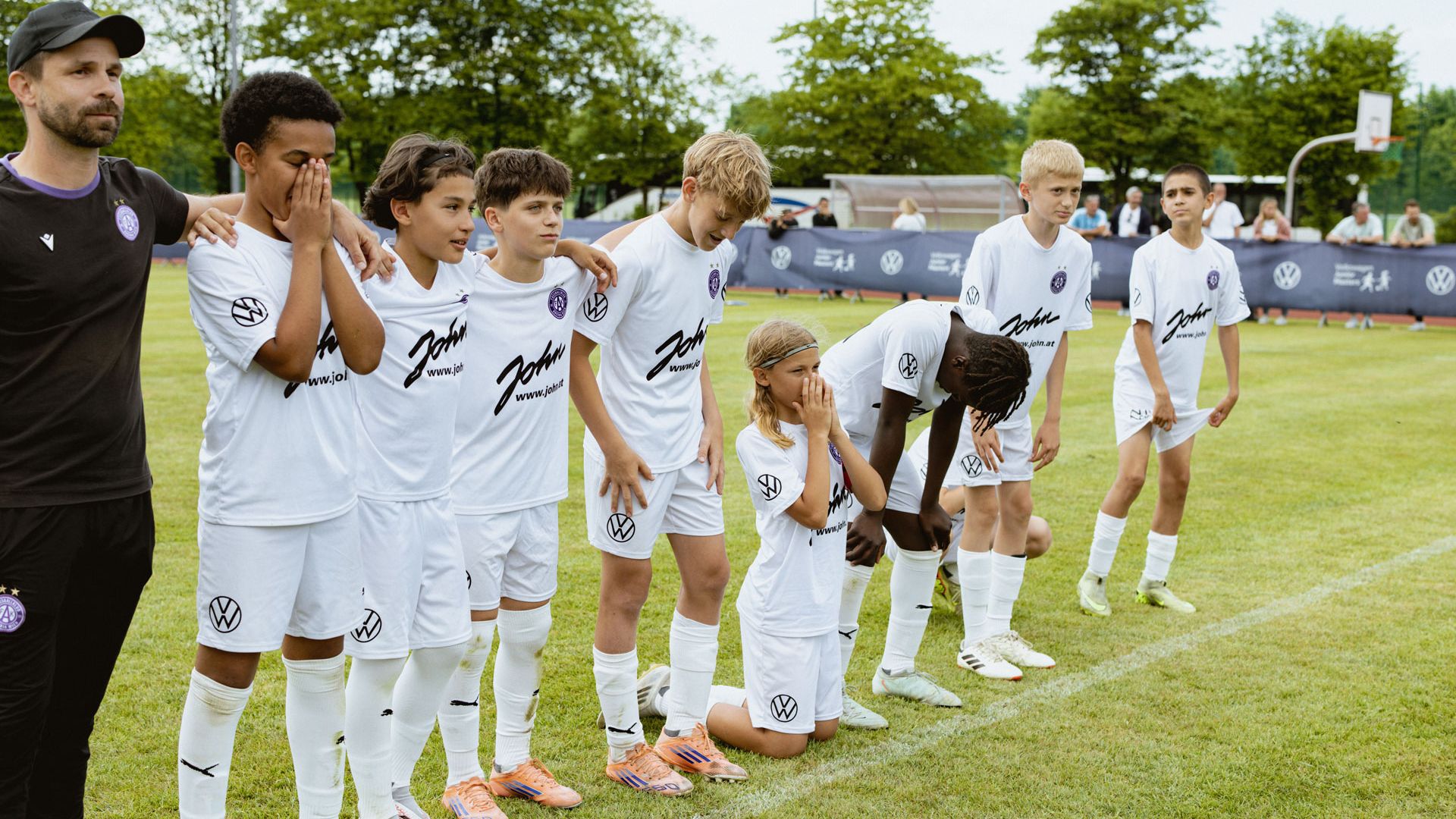Eine Jugendfußballmannschaft in weißen Trikots steht in einer Reihe auf dem Spielfeld, eine Spielerin kniet vorne mit gefalteten Händen. Die Kinder wirken konzentriert oder angespannt, als würden sie auf den Beginn oder das Ergebnis eines Spiels warten. Im Hintergrund sind Zuschauer, Bäume und ein einfacher Fußballplatz zu sehen.