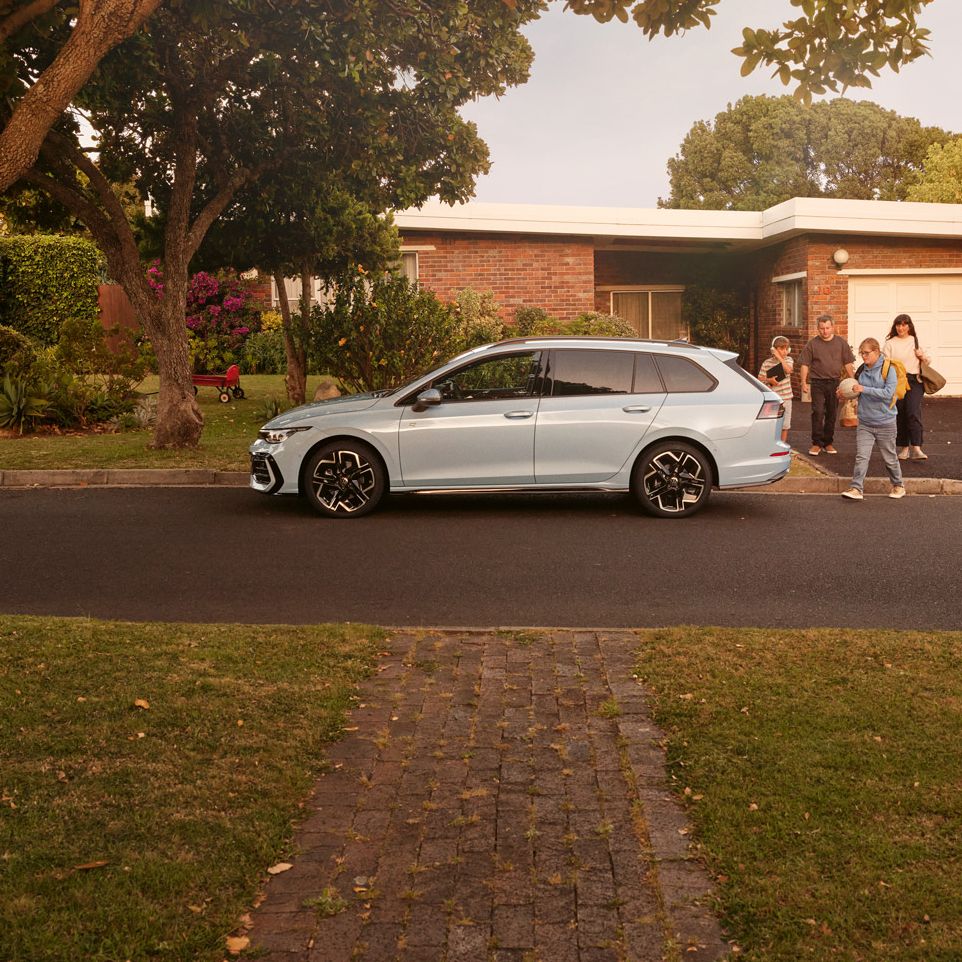 Ein weißer VW Golf Variant parkt vor einem Bungalow in einer Wohnstraße mit Bäumen. 