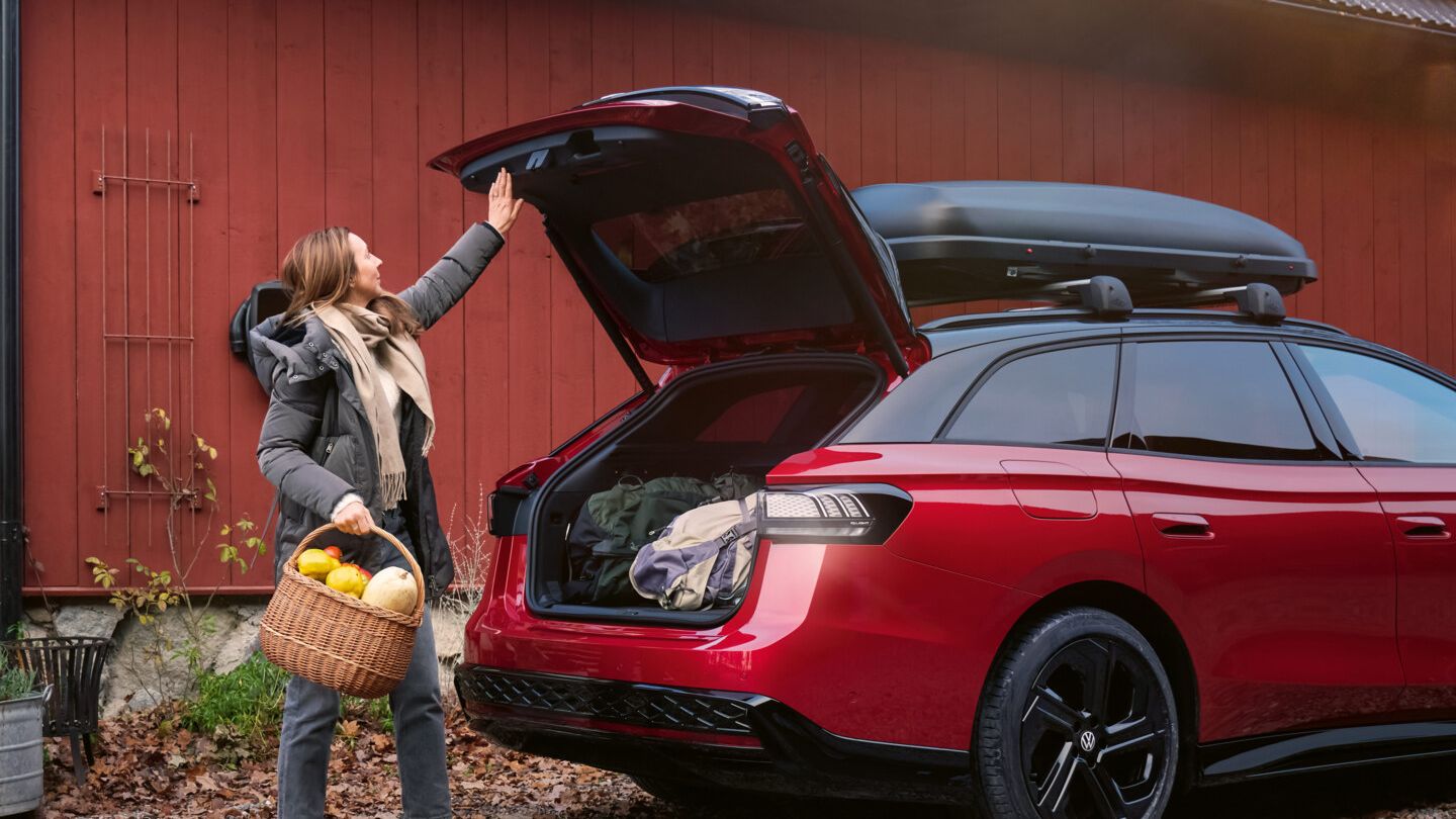 Eine Frau mit einem Korb voll Gemüse in der Hand steht vor der offenen Heckklappe eines roten VW ID.7 GTX Tourer.