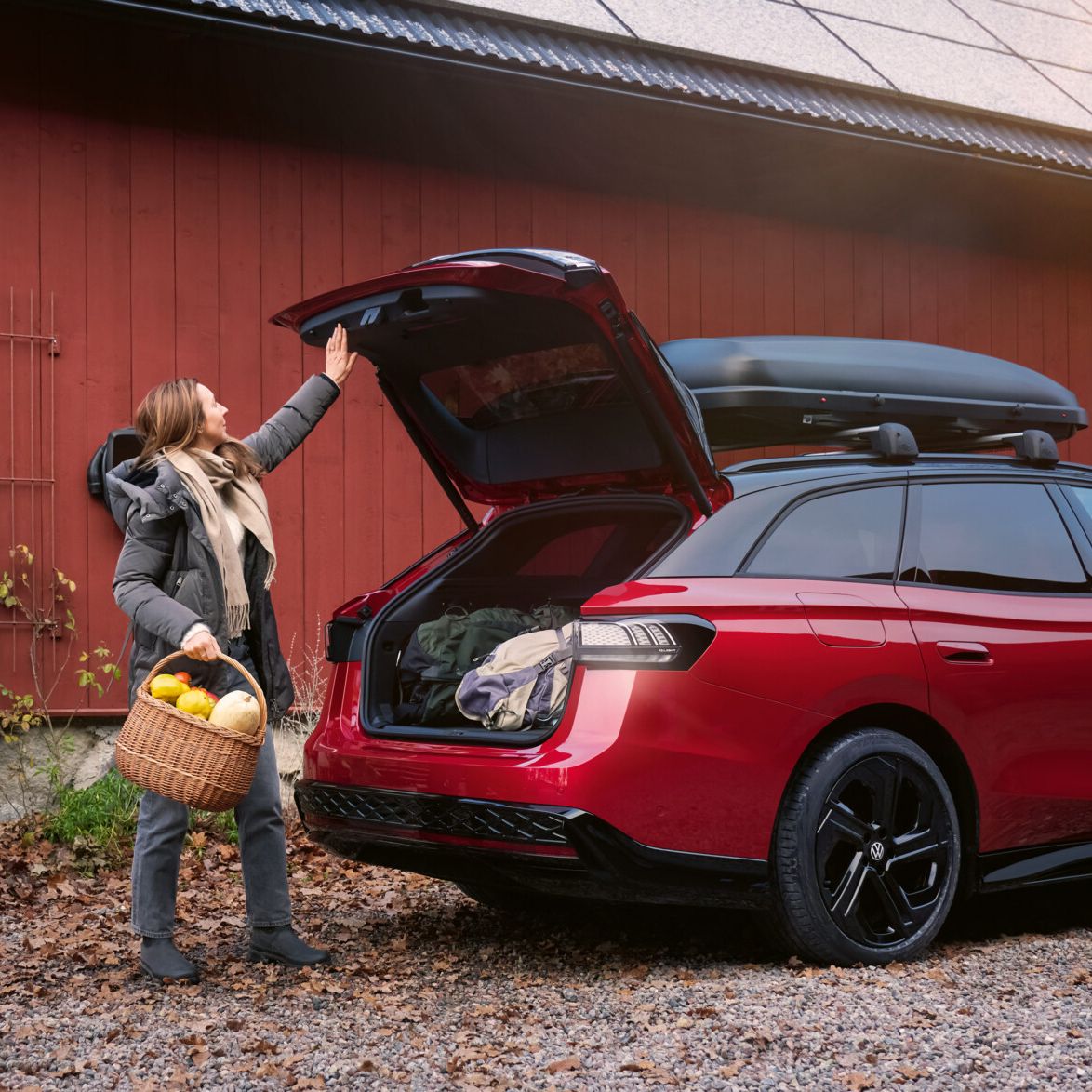 Eine Frau mit einem Korb voll Gemüse in der Hand steht vor der offenen Heckklappe eines roten VW ID.7 GTX Tourer.