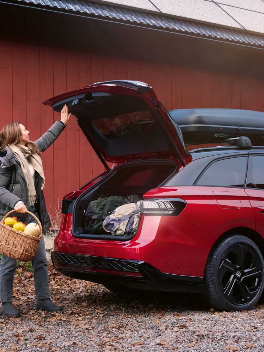 Eine Frau mit einem Korb voll Gemüse in der Hand steht vor der offenen Heckklappe eines roten VW ID.7 GTX Tourer.