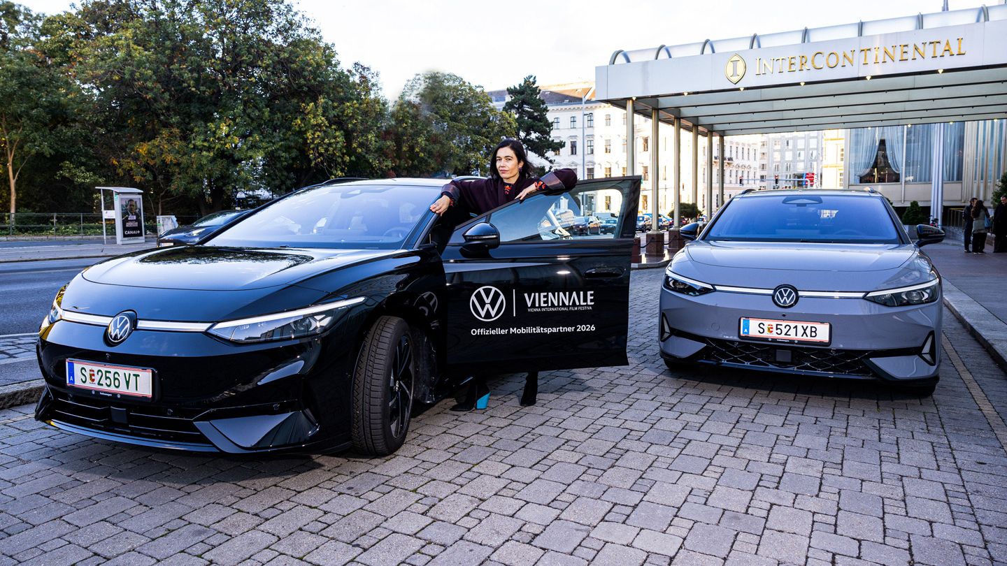 Eine Person steht vor dem Eingang des InterContinental Hotels neben einem schwarzen Volkswagen-Elektroauto und hält die geöffnete Fahrertür. Rechts daneben steht ein zweites, graues Elektroauto desselben Modells. Auf der Tür des schwarzen Wagens ist ein Logo der Viennale mit dem Hinweis „Offizieller Mobilitätspartner 2024“ zu sehen.