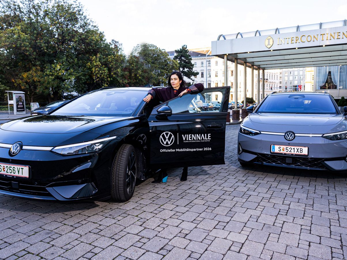 Eine Person steht vor dem Eingang des InterContinental Hotels neben einem schwarzen Volkswagen-Elektroauto und hält die geöffnete Fahrertür. Rechts daneben steht ein zweites, graues Elektroauto desselben Modells. Auf der Tür des schwarzen Wagens ist ein Logo der Viennale mit dem Hinweis „Offizieller Mobilitätspartner 2024“ zu sehen.