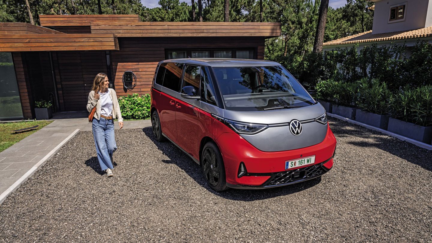 Eine Person steht neben einem roten Volkswagen ID. Buzz GTX, der in einer gepflasterten Einfahrt vor einem modernen Haus geparkt ist.