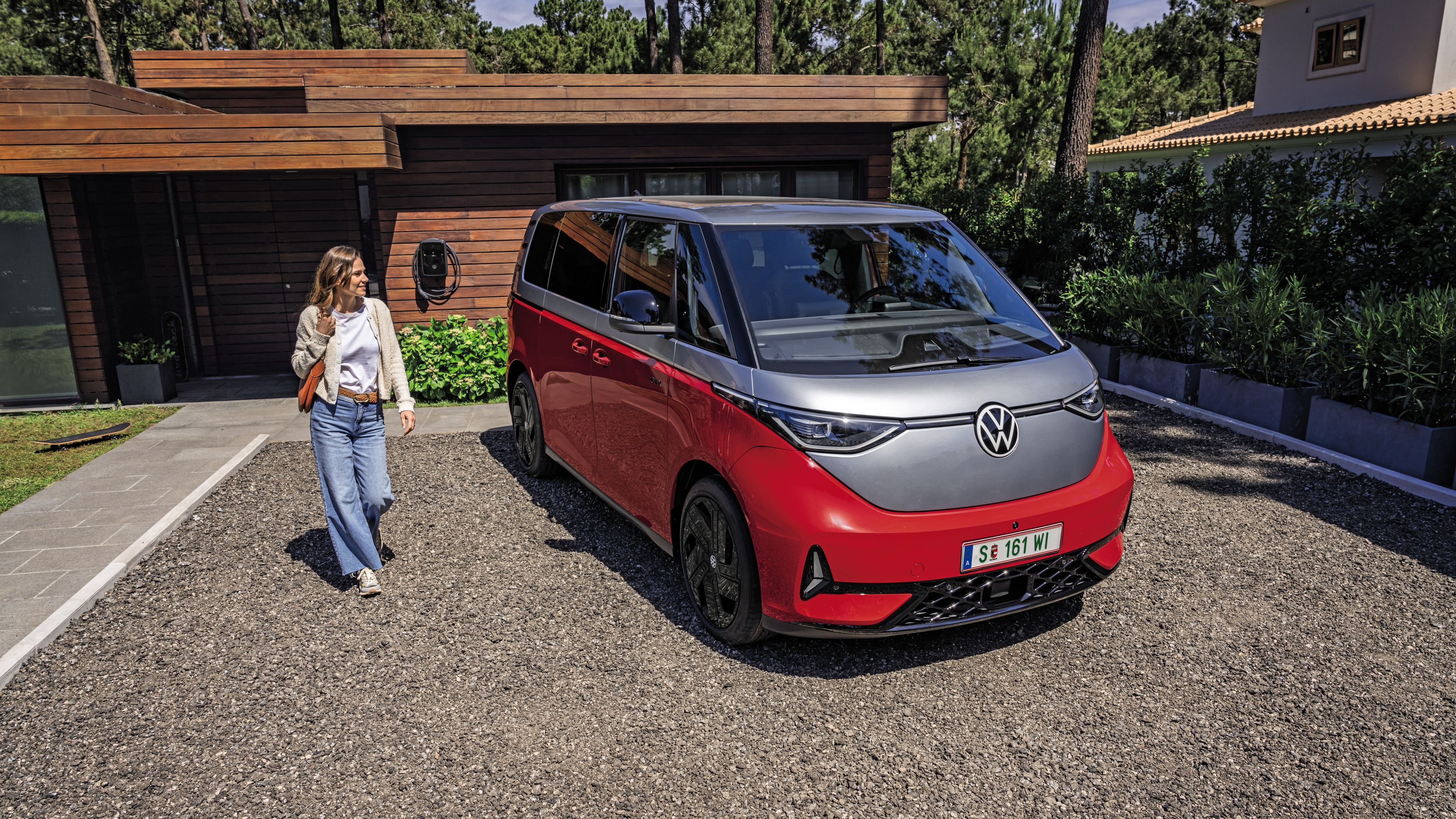 Eine Person steht neben einem roten Volkswagen ID. Buzz GTX, der in einer gepflasterten Einfahrt vor einem modernen Haus geparkt ist.