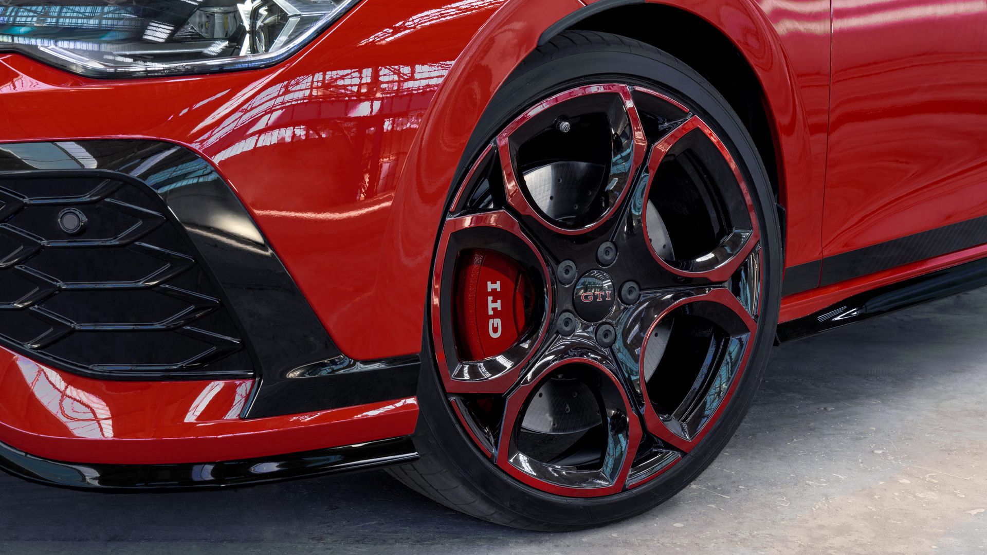 Nahaufnahme eines roten Golf GTI, das eine markante schwarze und rote Felge mit dem GTI-Logo zeigt. Die Frontschürze ist teils sichtbar und hat eine Wabenstruktur. Das Auto steht in einer hellen Umgebung mit viel natürlichem Licht.