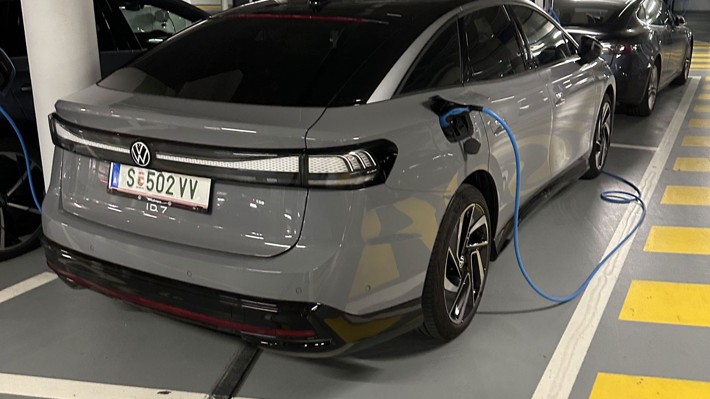 Ein grauer ID.7 von Volkswagen wird in einer Tiefgarage an einer Ladesäule aufgeladen. Das KFZ-Kennzeichen zeigt an, dass das Auto aus Österreich stammt. Rechts im Hintergrund befindet sich ein weiteres Fahrzeug.