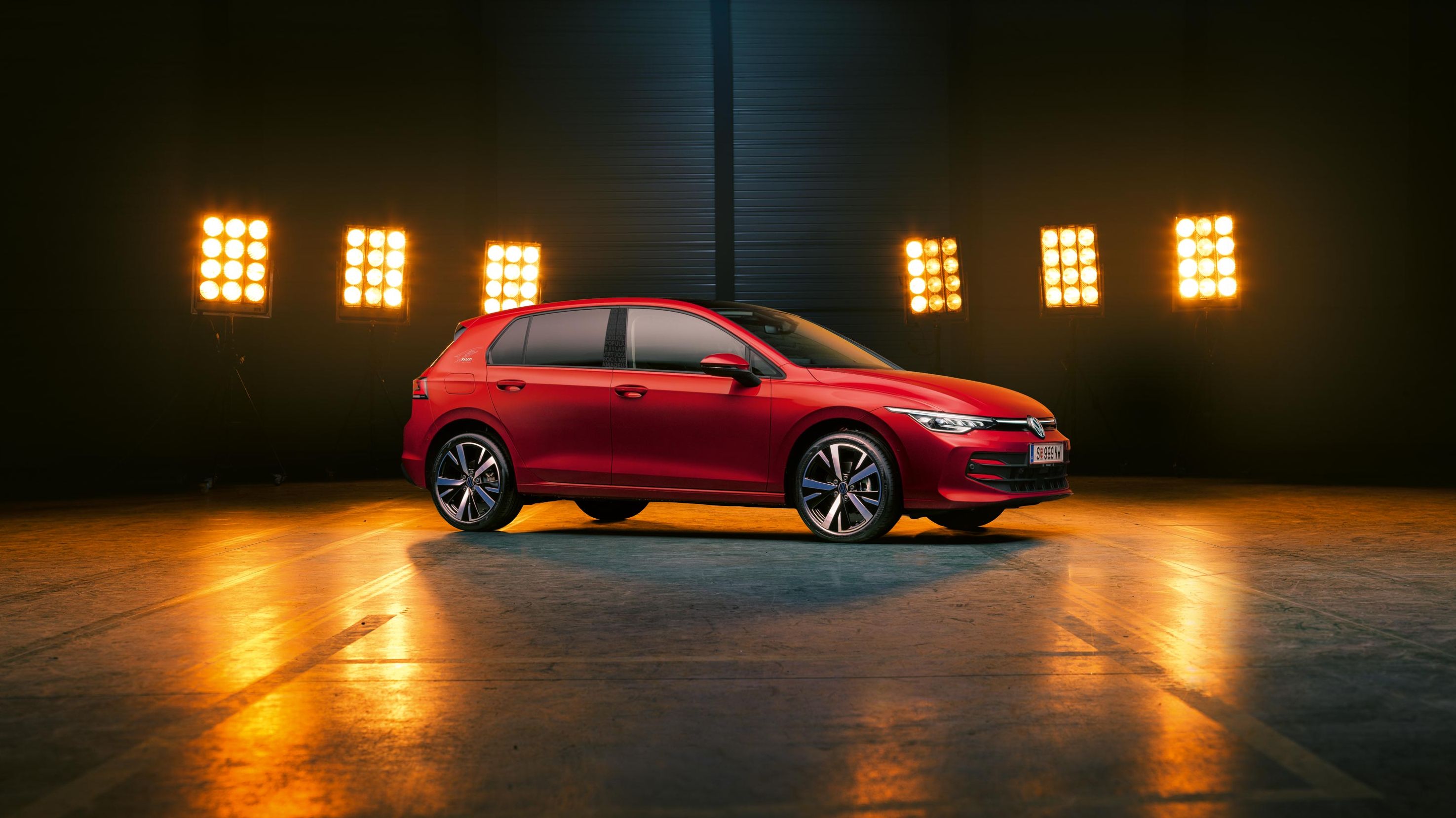Seitenansicht auf einen roten VW Golf Falco Edition, der in einer leeren Halle steht, dahinter wird er von Scheinwerfern beleuchtet.