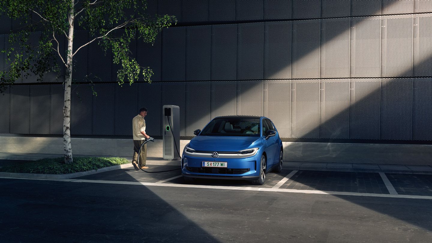 Ein blauer ID. Polo steht auf einem Parkplatz neben einer Ladesäule. Im Hintergrund sind ein Gebäude und ein Baum zu sehen.