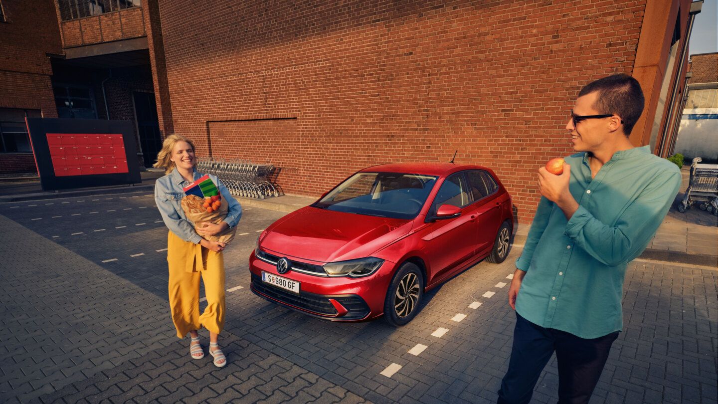 Ein roter VW Polo steht auf einem Parkplatz vor einem Supermarkt, ein lachendes Paar steht mit Einkäufen daneben.