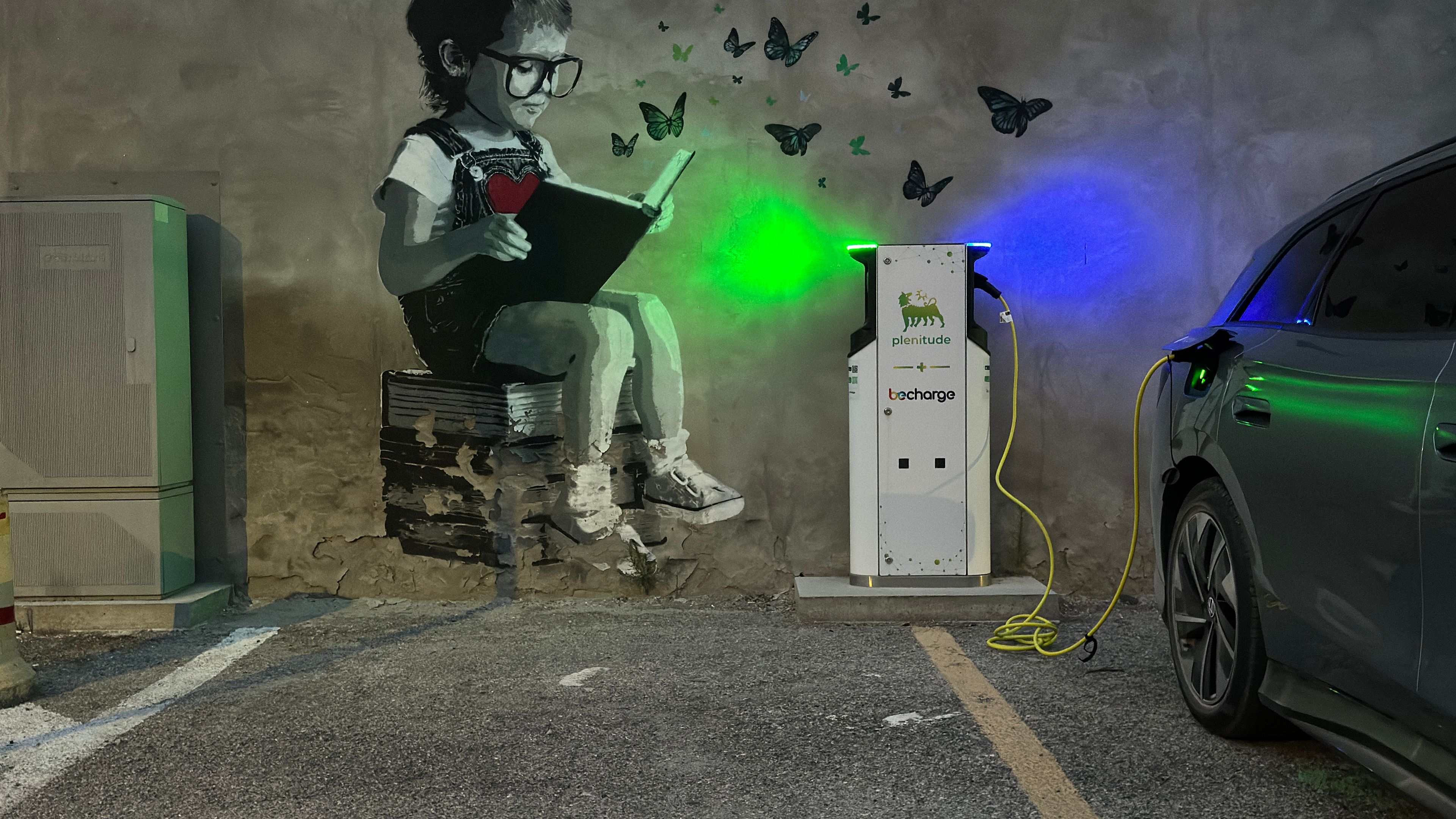 Ein graffitierter Parkplatz zeigt ein Kind, das auf einem Buch sitzt und liest, während Schmetterlinge umherfliegen. Daneben lädt ein Elektroauto an einer Ladestation. Im Hintergrund ist eine Wand aus Beton.