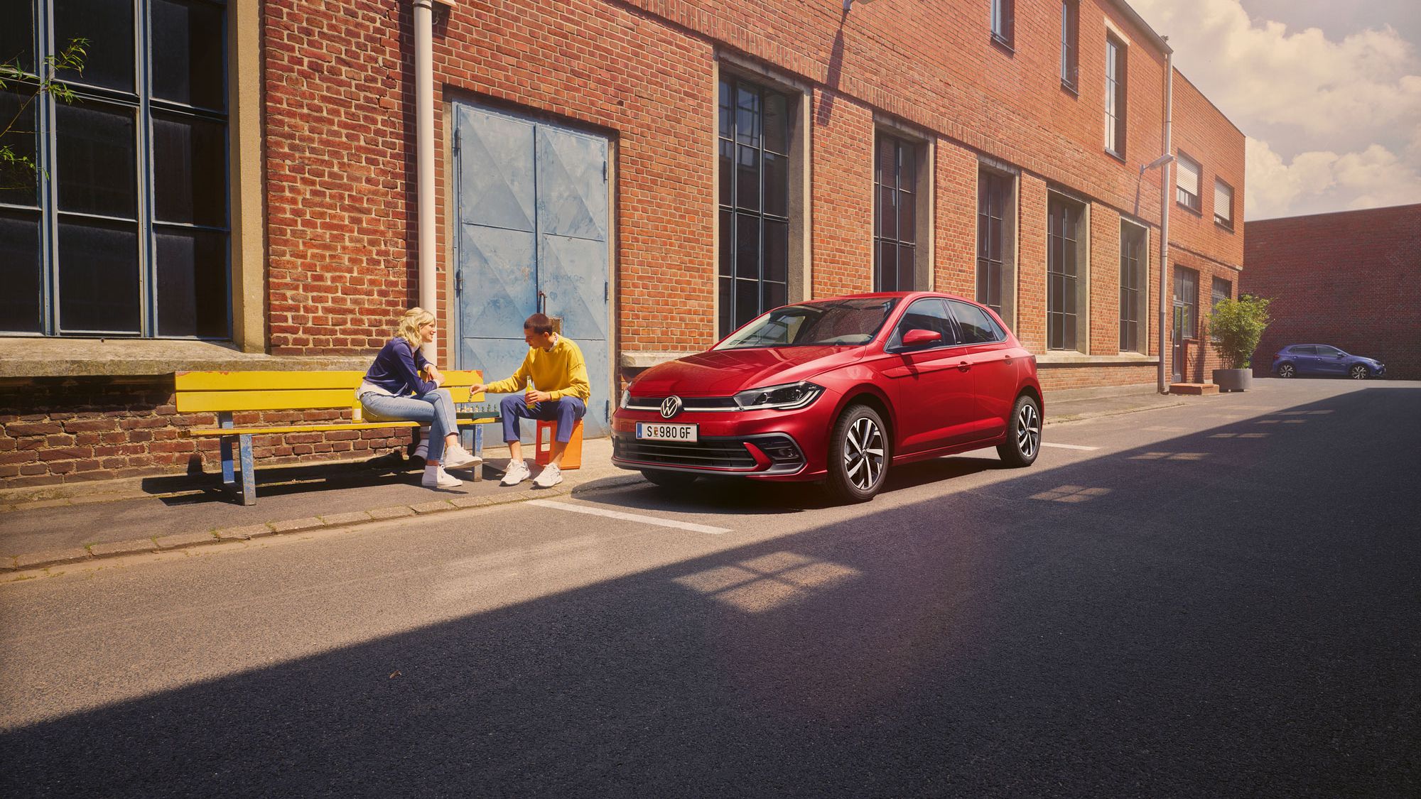 Zwei Personen spielen Schach vor einem roten VW Polo Style, der vor einem Fabrikgebäude parkt.
