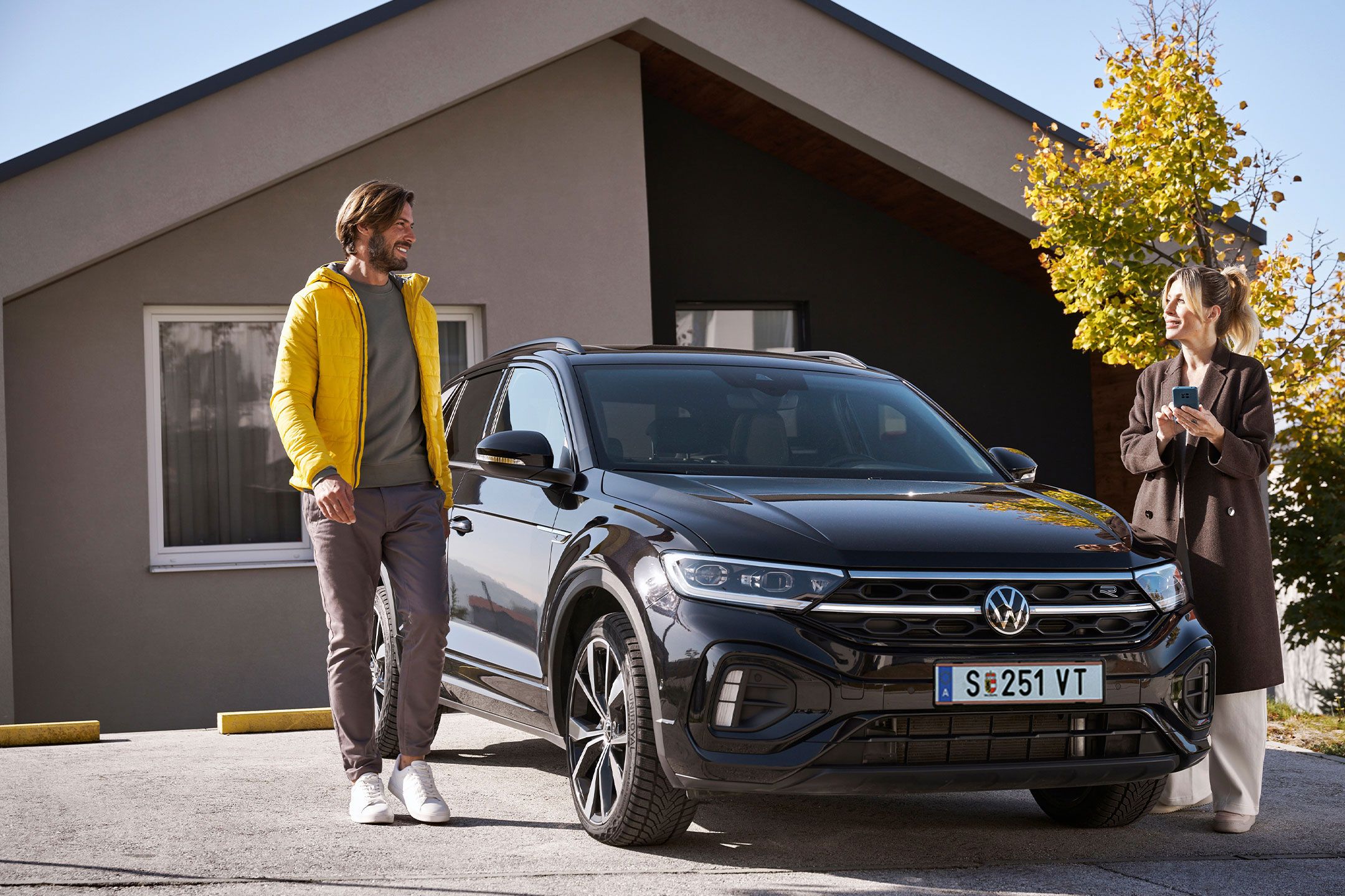 Mann und Frau steht mit Smartphone neben einem geparkten T-Roc vor einem Haus.