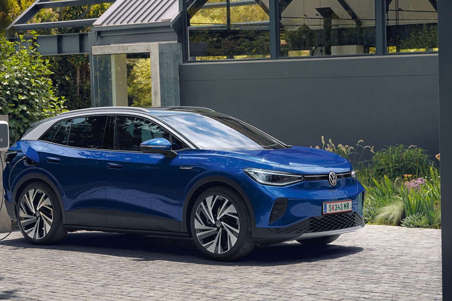 Ein blauer VW ID.4 parkt seitlich vor einem grauen, modernen Haus und lädt an der ID. Charger Wallbox. Im Hintergrund viel grün.
