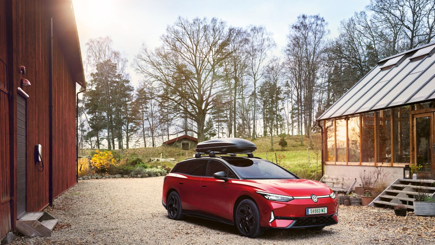 Roter VW ID.7 GTX Tourermit Dachbox parkt zwischen zwei Holzhäusern, im Hintergrund ein Wald.