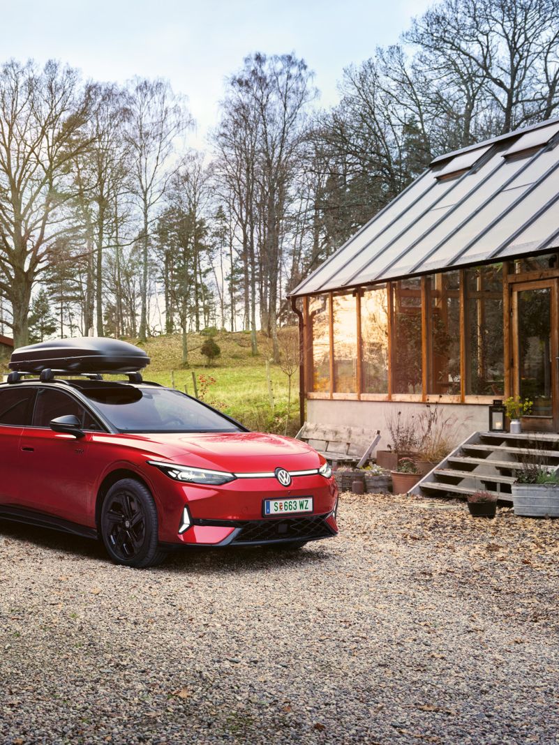 Roter VW ID.7 GTX Tourermit Dachbox parkt zwischen zwei Holzhäusern, im Hintergrund ein Wald.