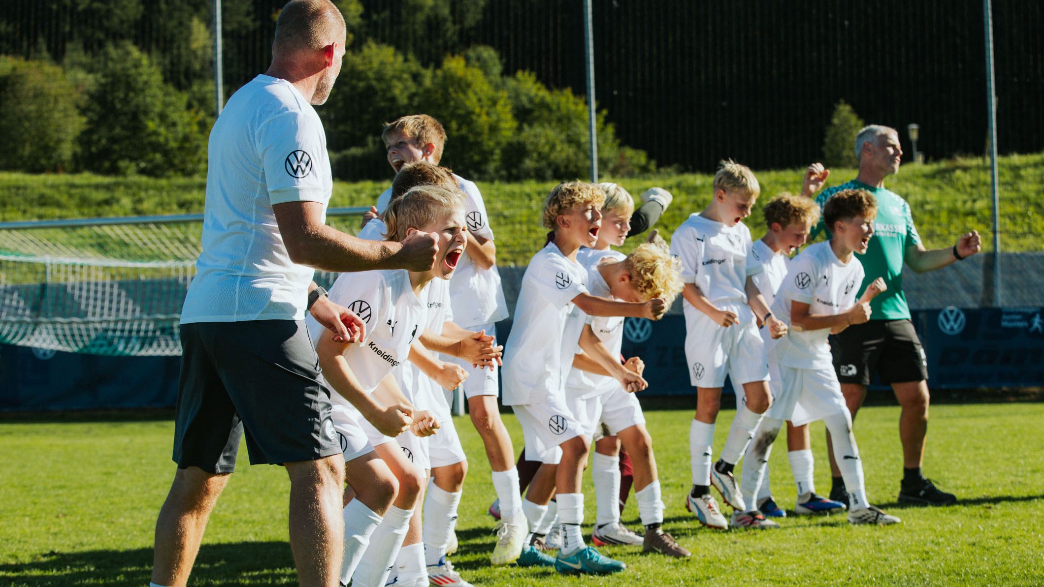 Eine Gruppe junger Fußballspieler in weißen Trikots jubelt ausgelassen auf einem sonnigen Spielfeld. Zwei erwachsene Trainer stehen links und rechts bei der Mannschaft und feuern die Kinder an. Im Hintergrund sind ein grüner Hang und Bäume zu sehen.
