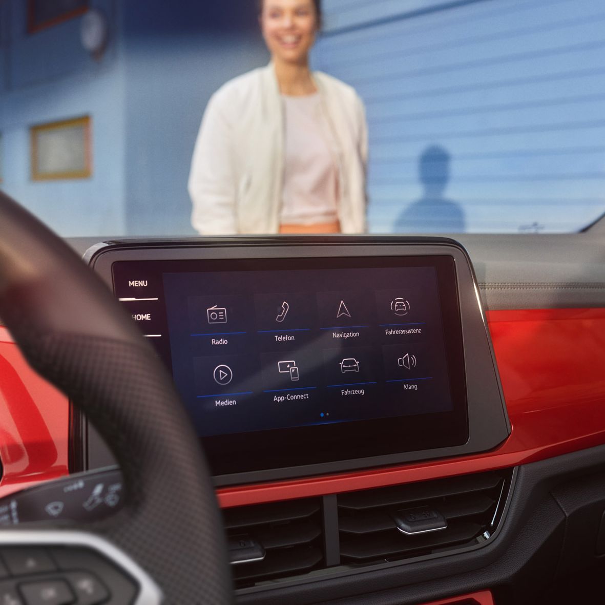 Das Bild zeigt das Innenleben eines VW T-Roc Cabrios mit einem modernen Infotainment-System. Im Hintergrund steht eine Person vor einer blauen Wand. Die Armaturenbrettdetails, wie rote Akzente, sind sichtbar.