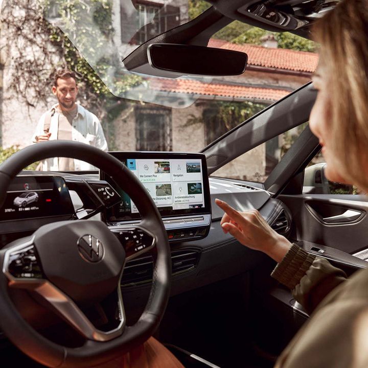 Eine Frau sitzt in einem VW und bedient den großen Touchscreen des Infotainmentsystems. Im Hintergrund steht ein Mann vor einem Haus. 