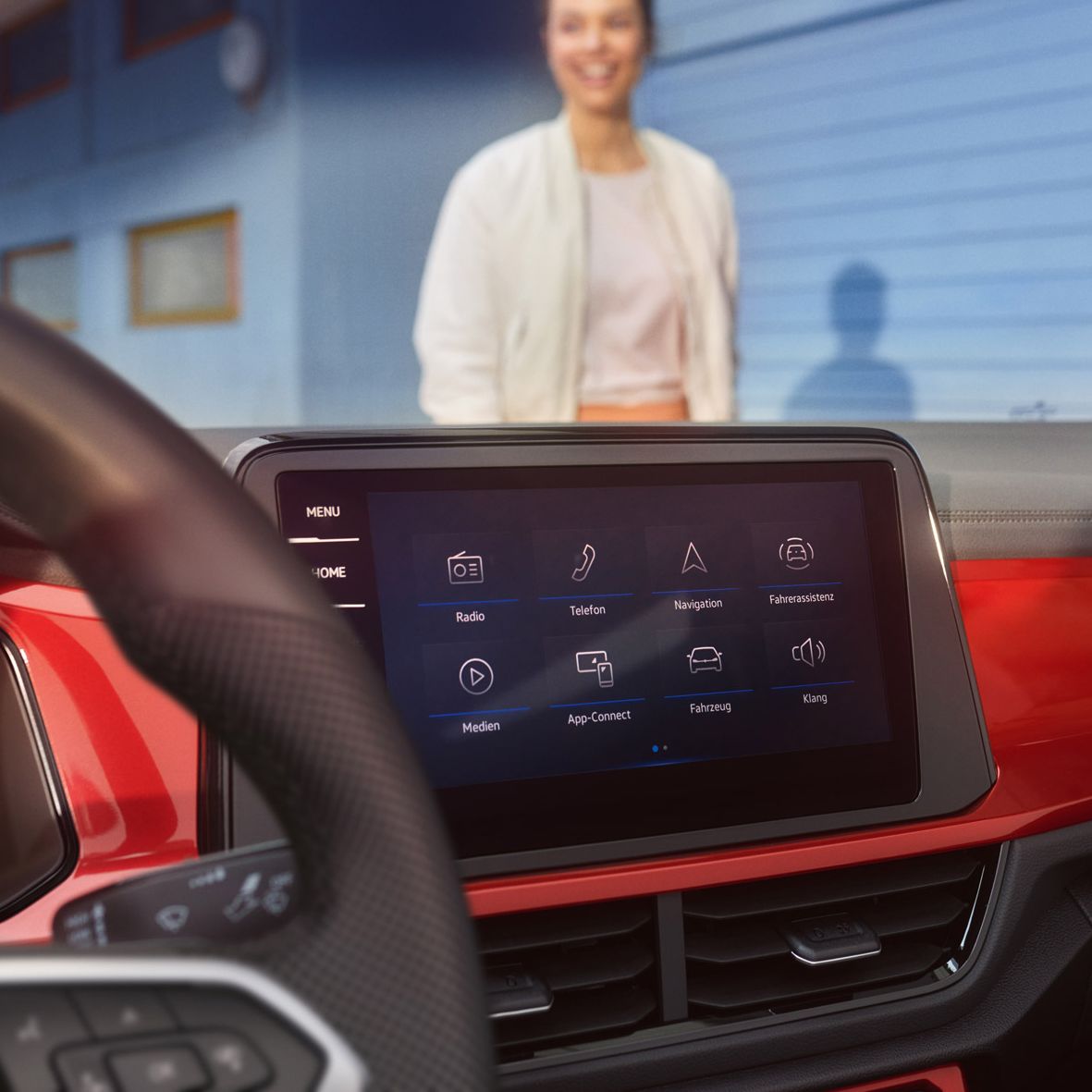 Das Bild zeigt das Innenleben eines VW T-Roc Cabrios mit einem modernen Infotainment-System. Im Hintergrund steht eine Person vor einer blauen Wand. Die Armaturenbrettdetails, wie rote Akzente, sind sichtbar.