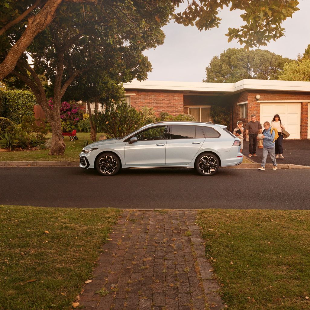 Ein weißer VW Golf Variant parkt vor einem Bungalow in einer Wohnstraße mit Bäumen. 
