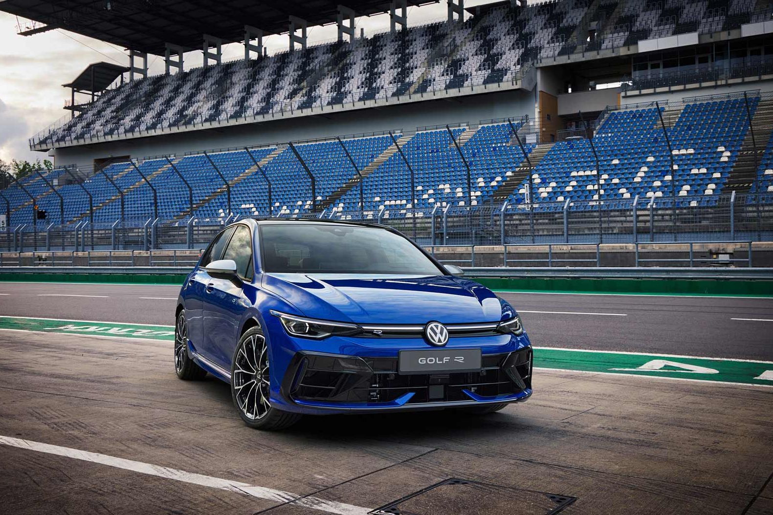 Ein blauer Volkswagen Golf R steht auf einer Rennstrecke vor einer leeren Tribüne. Das Fahrzeug ist sportlich designiert und hat eine dynamische Frontansicht. Im Hintergrund sind graue und blaue Sitze zu sehen.
