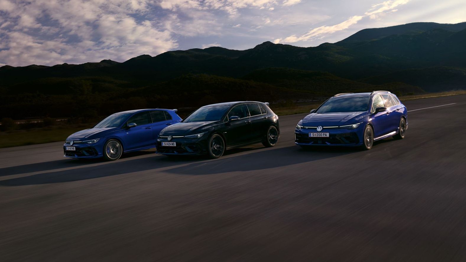 Drei VW Golf R fahren nebeneinander auf einer Straße bei Sonnenuntergang. Die Himmel zeigt einige Wolken und im Hintergrund sind Berge sichtbar. Die Farben der Autos sind blau, schwarz und dunkelblau.