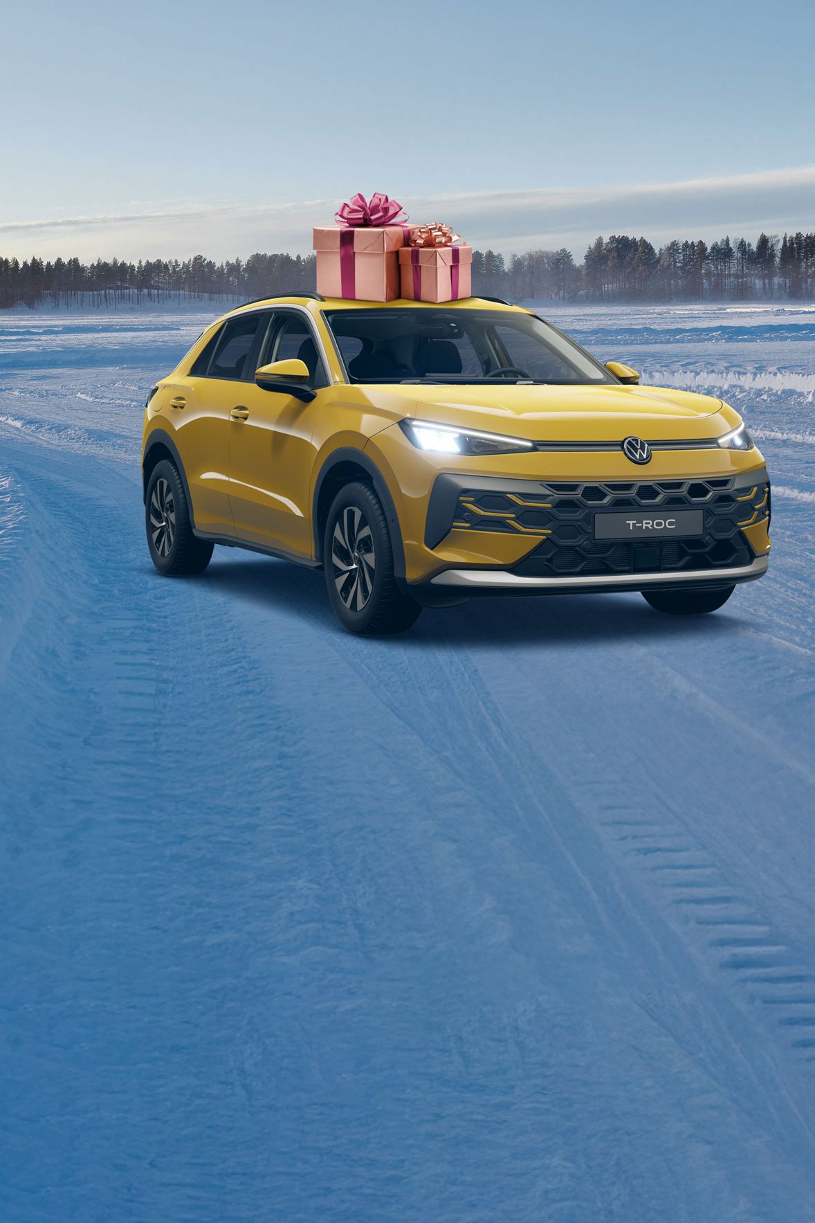 Gelber Volkswagen T-Roc fährt auf schneebedecktem Gelände. Auf dem Dach des Autos befinden sich zwei Geschenkkartons mit Schleifen. Im Hintergrund ist eine bewaldete Landschaft zu sehen.