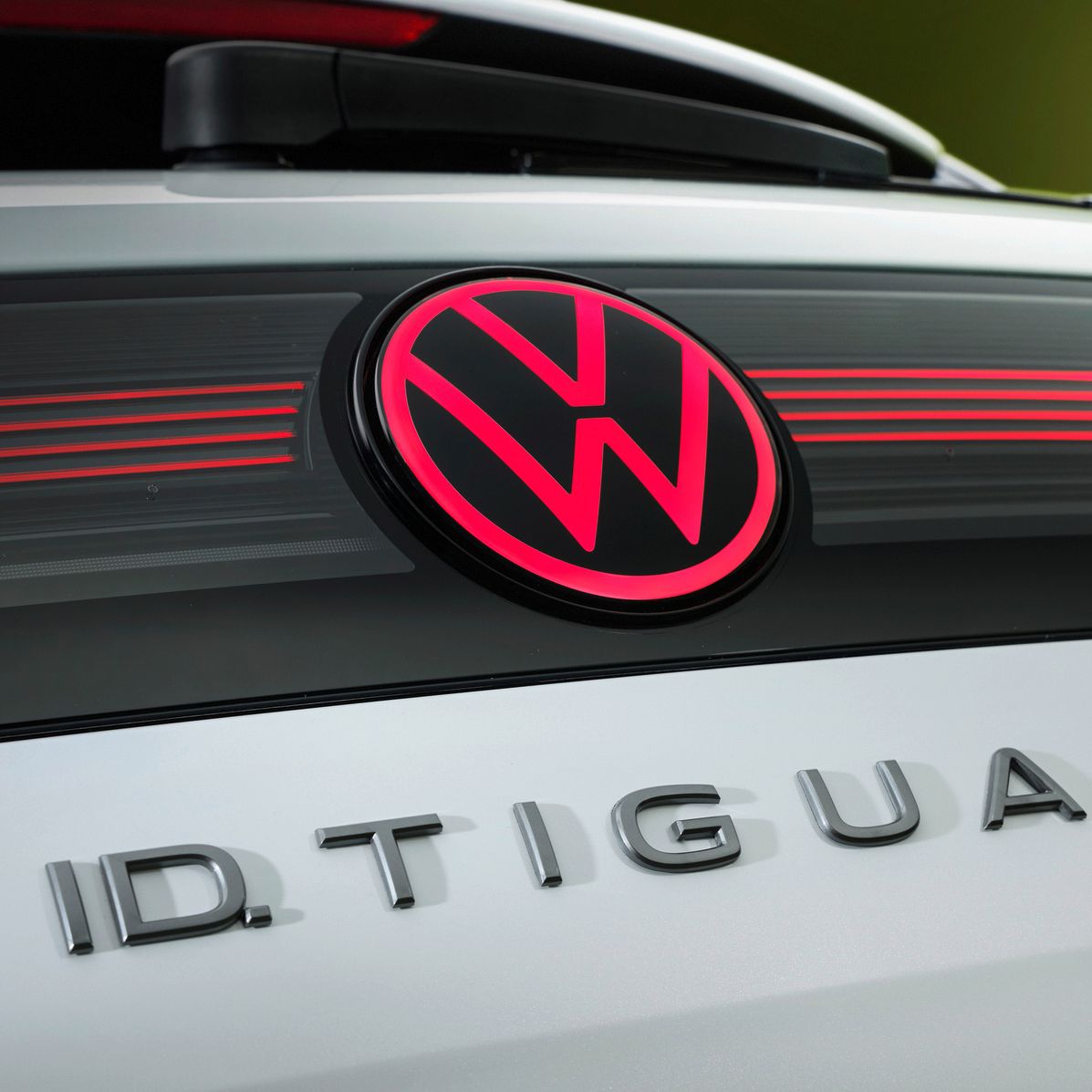 Nahaufnahme des Hecks eines weißen Volkswagen ID. Tiguan mit zentral platziertem, rot leuchtendem VW-Logo. Darunter ist in silbernen Buchstaben der Modellname „ID. TIGUAN“ auf der Heckklappe zu sehen. Im Hintergrund ist ein unscharfer, grünlicher Farbverlauf erkennbar.