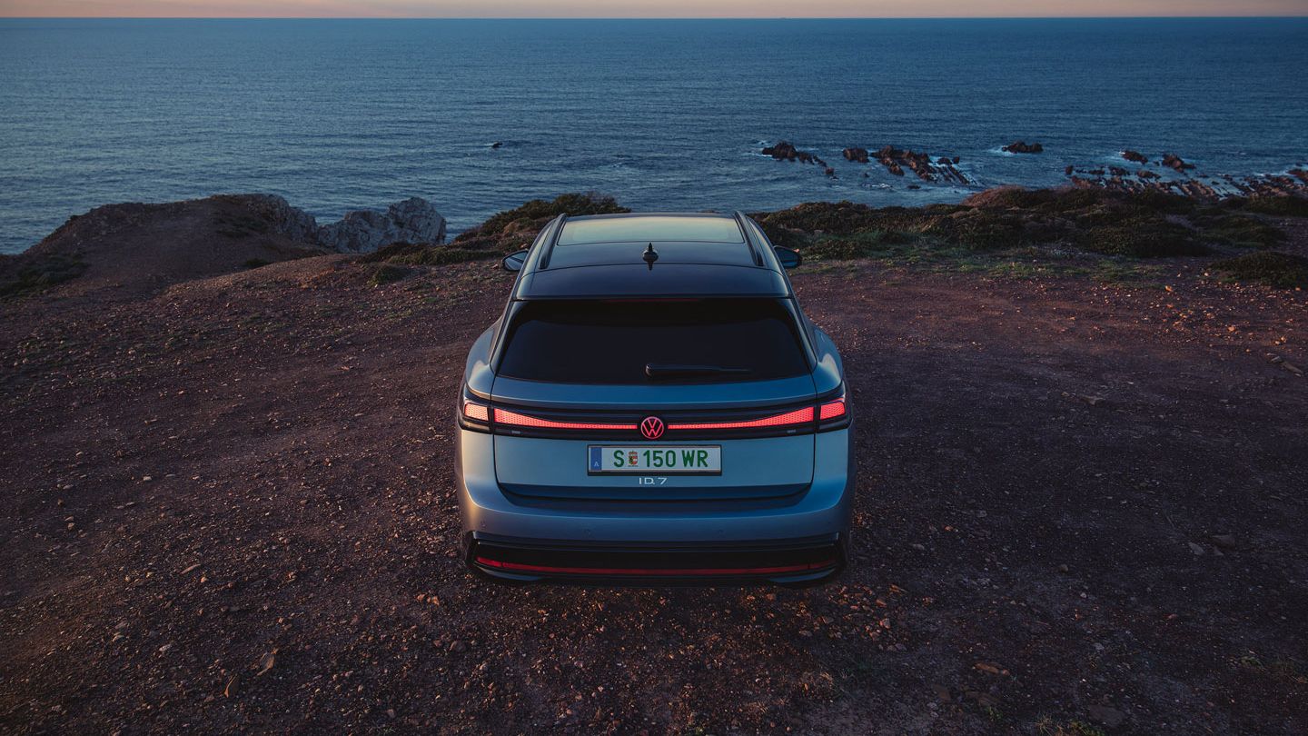 Blauer VW ID.7 Tourer in Heckansicht steht auf einer Klippe am Meer, die Heck-Lichtsignatur ist angeschaltet.