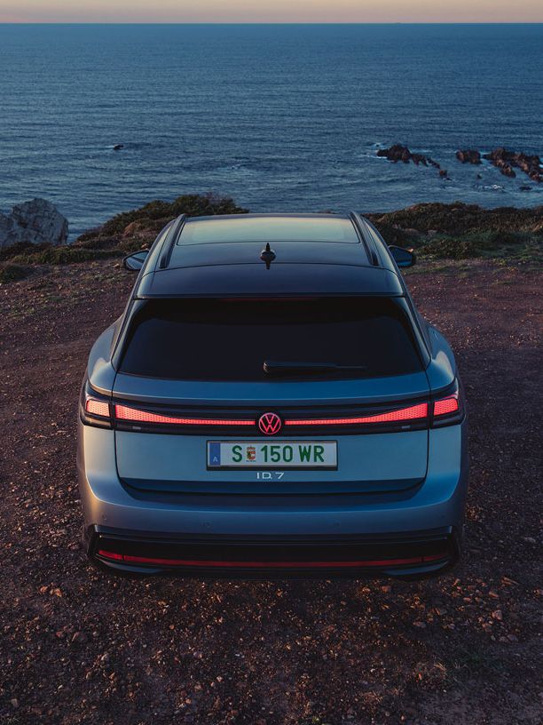 Blauer VW ID.7 Tourer in Heckansicht steht auf einer Klippe am Meer, die Heck-Lichtsignatur ist angeschaltet.
