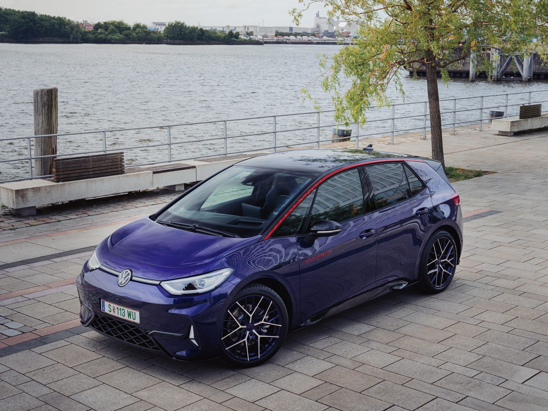 Ein blauer Volkswagen ID.3 GTX steht auf einem gepflasterten Platz am Ufer eines Gewässers. Der Hintergrund zeigt Bäume und industrielle Strukturen. Das Auto hat schwarze Felgen und rote Akzente.