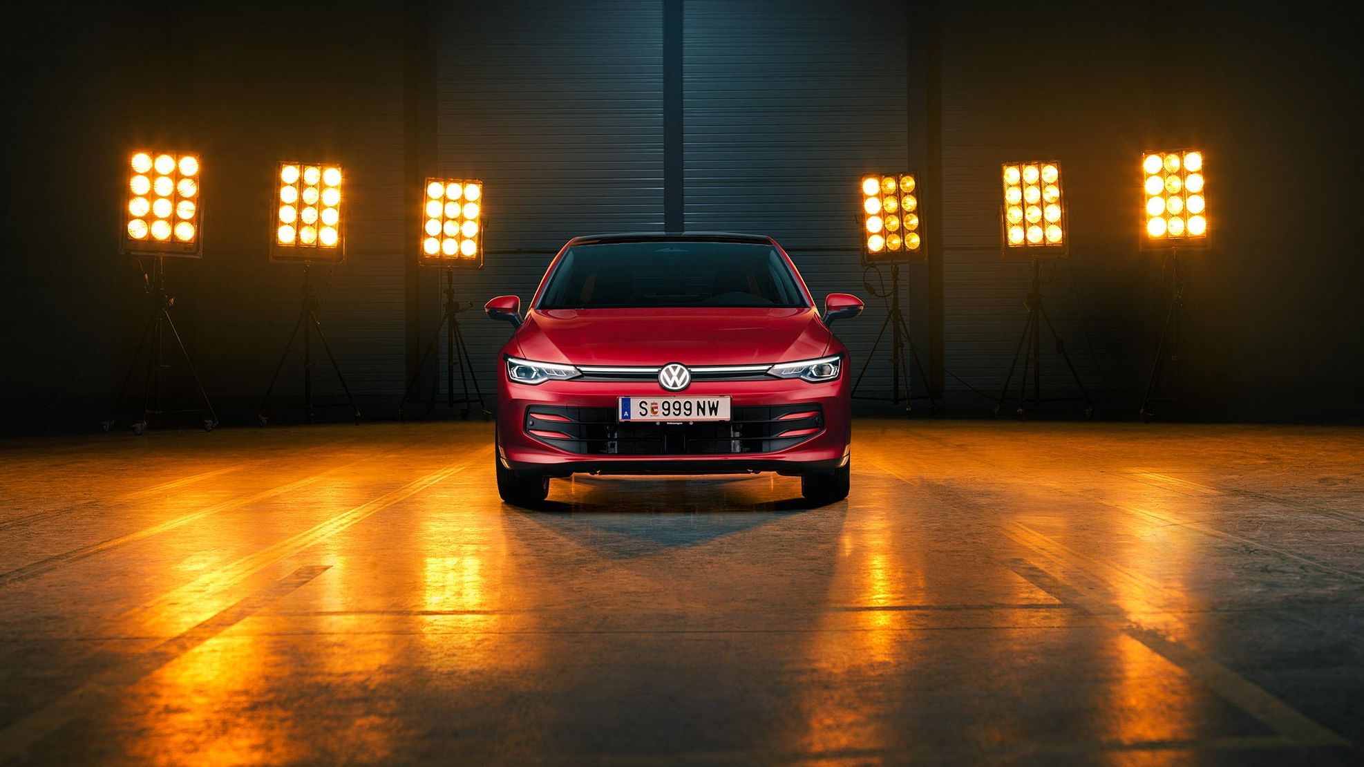 Ein roter VW Golf Falco Edition in Frontalansicht steht mittig im Bild in einer leeren Halle, Bühnenlichter strahlen das Fahrzeug von hinten an.