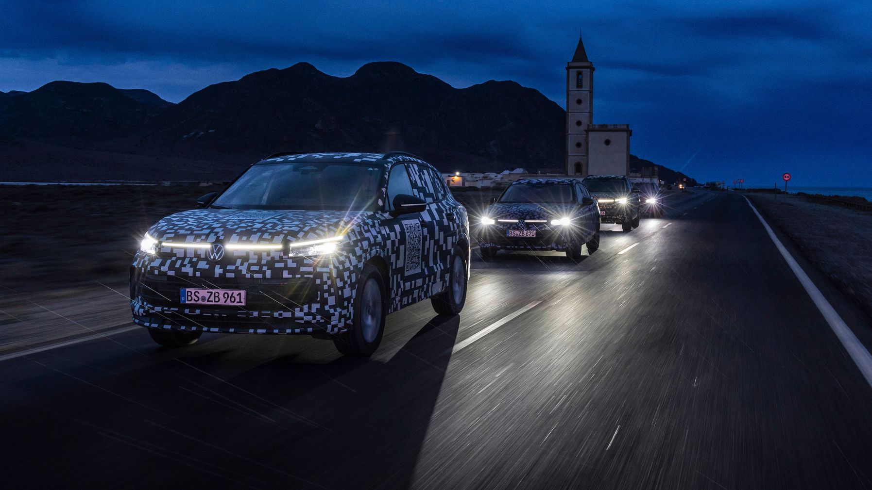 Mehrere getarnte Autos fahren auf einer Straße in der Dämmerung. Im Hintergrund sind Berglandschaften und eine Kirche sichtbar.