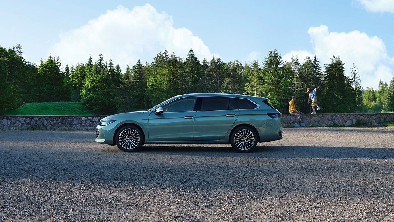 Ein grüner VW Passat B9 in der Seitenansicht, im Hintergrund zwei Menschen und Wald