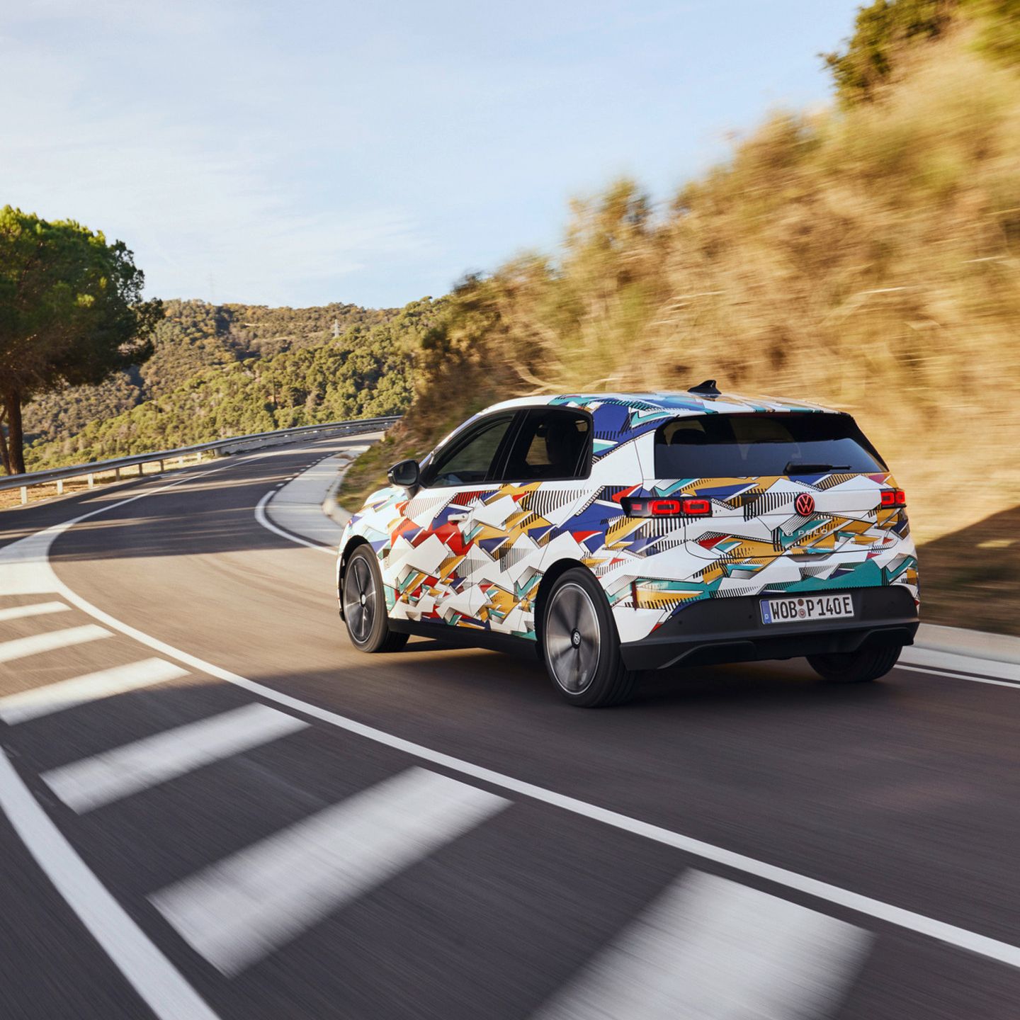 Ein ID. Polo mit bunter Lackierung fährt auf einer kurvenreichen Straße durch eine hügelige Landschaft. Auf der rechten Straßenseite sind Bäume und Büsche zu sehen. Der Himmel ist klar.