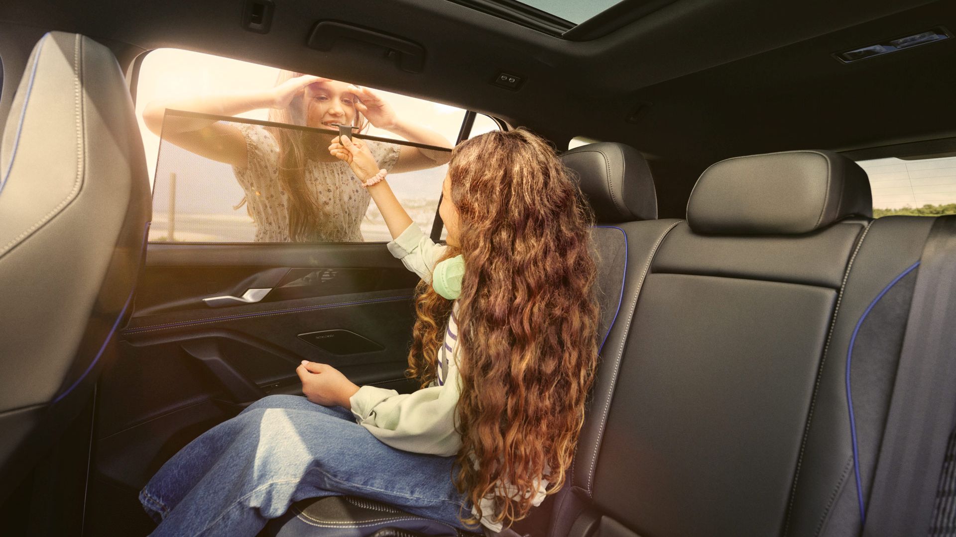 Ein Mädchen sitzt auf dem Rücksitz eines VW Tayron und öffnet das Sonnenrollo, eine Frau schaut durch das Fenster hinein.