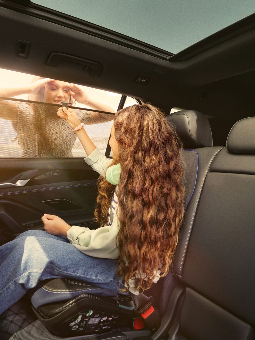 Ein Mädchen sitzt auf dem Rücksitz eines VW Tayron und öffnet das Sonnenrollo, eine Frau schaut durch das Fenster hinein.