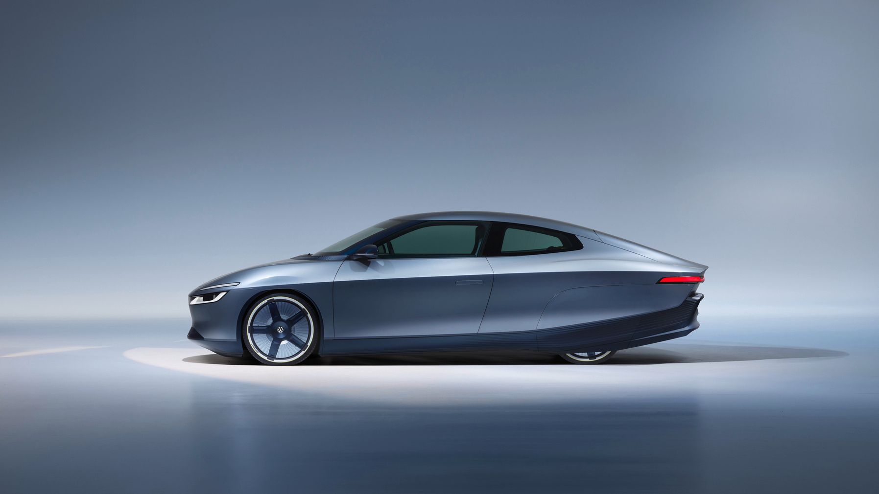 Seitlich aufgenommenes, futuristisches Elektroauto in dunkelgrauer Metallic-Lackierung vor neutralem, blau-grauem Studiohintergrund. Die Karosserie ist sehr flach und aerodynamisch mit glatten Flächen, verdeckten Türgriffen und schmalen Leuchten. Die Felgen haben ein nahezu geschlossenes, minimalistisches Design.
