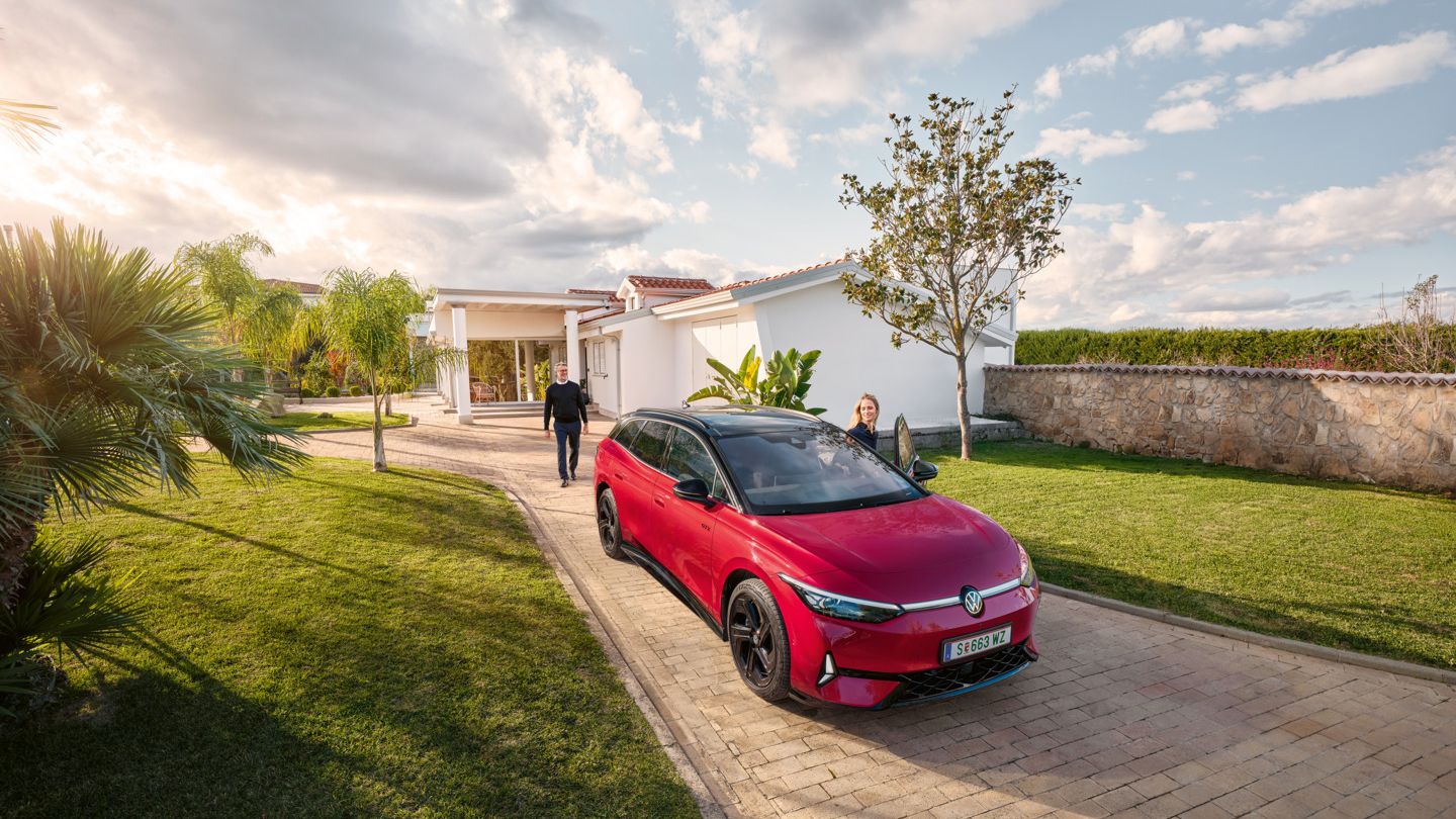 Ein roter VW ID.7 GTX parkt vor einer mediterranen Villa mit großem Garten, eine Frau steigt ein.