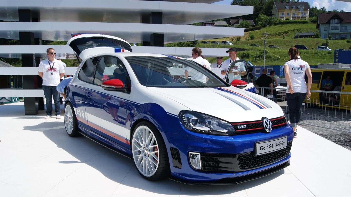 Ein weißer Volkswagen Golf GTI mit blauen und roten Rennstreifen steht auf einem Ausstellungsbereich. Um das Auto herum stehen Menschen und im Hintergrund sind Gebäude und ein grüner Hügel zu sehen.