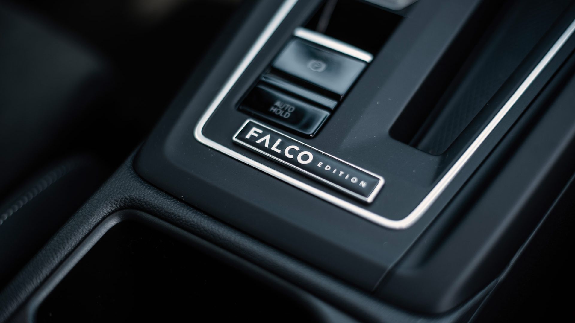 Nahaufnahme einer Mittelkonsole im Auto mit dem Schriftzug „FALCO EDITION“ auf einer dekorativen Plakette. Daneben sind Tasten und Bedienelemente zu sehen, darunter eine mit der Beschriftung „AUTO HOLD“. Die Oberfläche wirkt modern und hochwertig mit dunklen Materialien und klaren Linien.