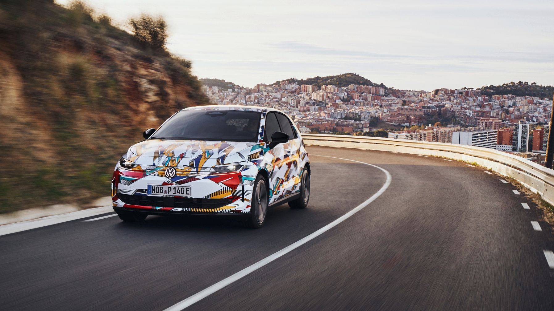 VW ID. Polo mit bunter Folierung fährt auf einer kurvenreichen Straße, im Hintergrund sind eine Stadt und Hügel zu sehen.