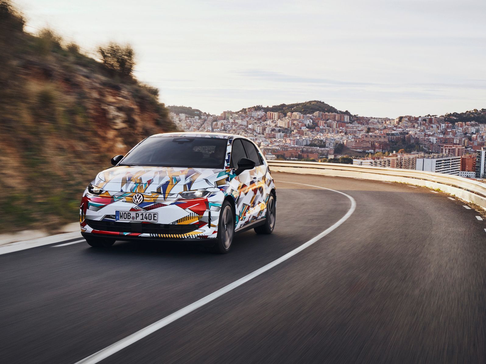 VW ID. Polo mit bunter Folierung fährt auf einer kurvenreichen Straße, im Hintergrund sind eine Stadt und Hügel zu sehen.