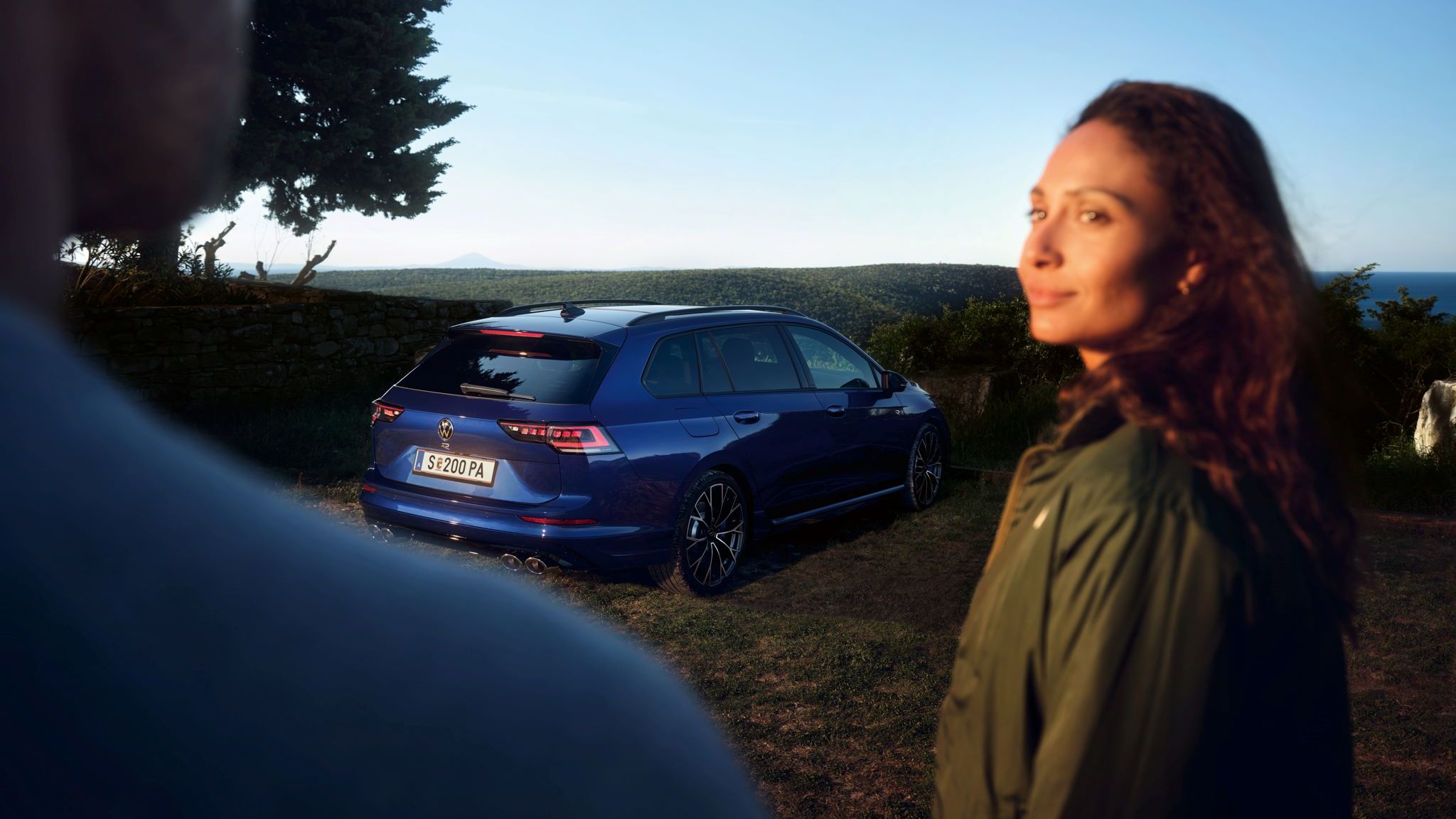 Frau und Mann im Vordergrund, im Hintergrund geparkter Golf Variant R in Heckansicht. 